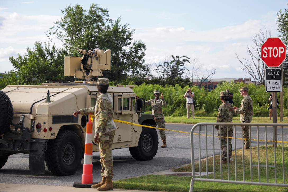 Four soldiers stand near a parked Humvee where two roads meet, with a stop sign, an orange cone, a security gate and two photographers also visible, along with green vegetation in the background.