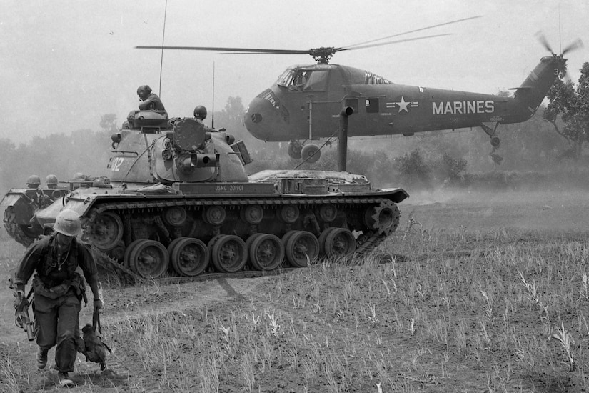 A helicopter hovers within feet of a field, with a military tank nearby. A person in a military uniform moves away from the tank.