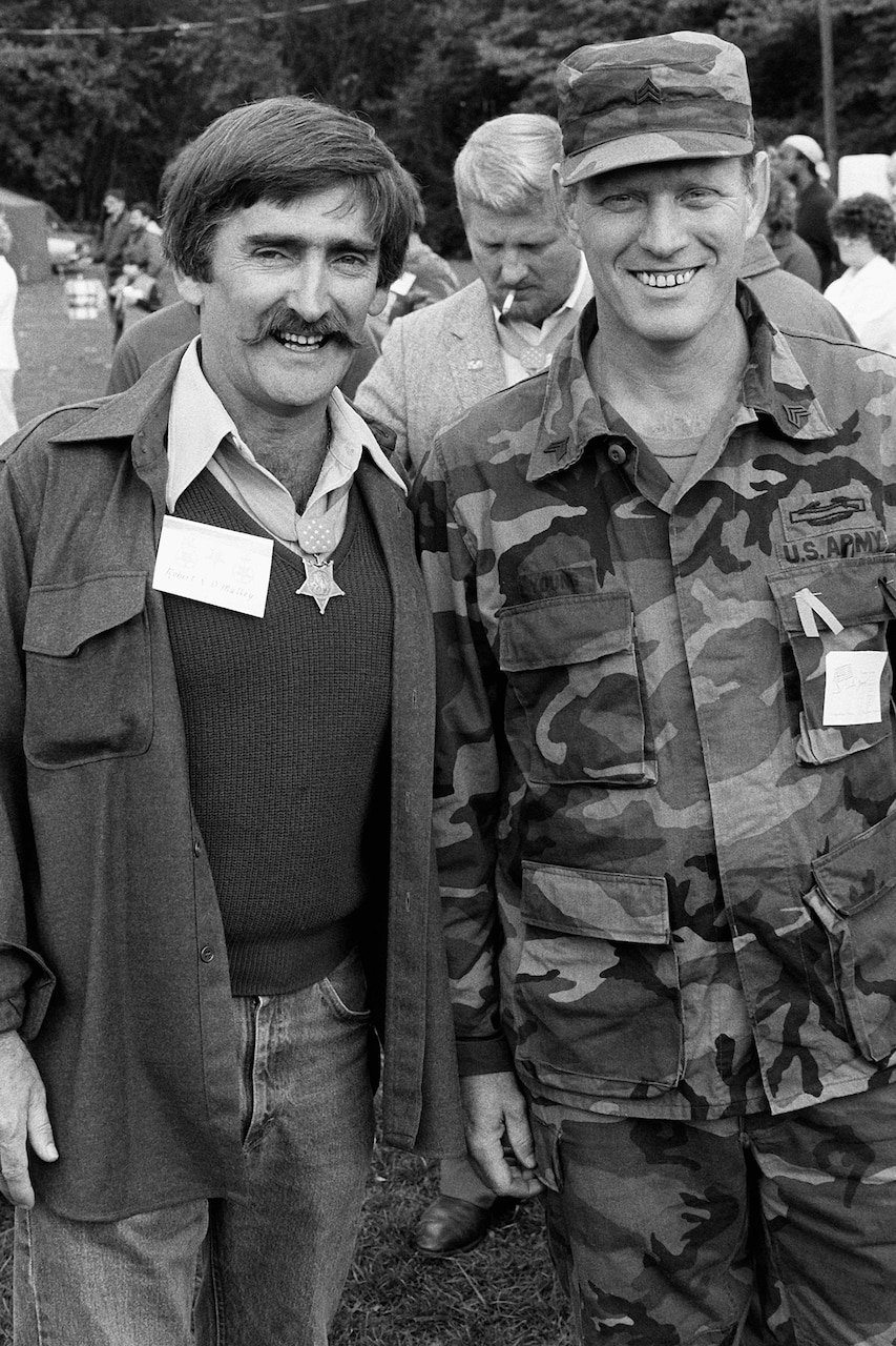 A person in a camouflage uniform and another person in casual attire wearing a medal around the neck stand beside each other and smile.
