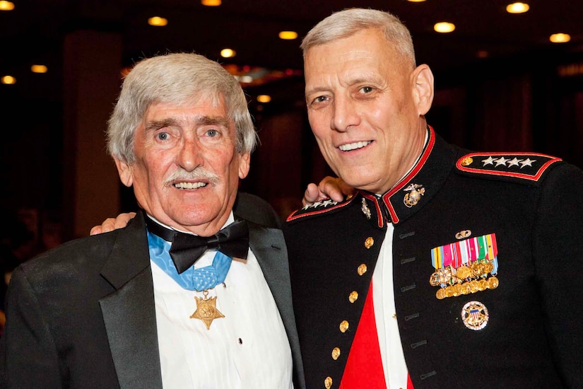 A person in a tuxedo and wearing a medal around the neck stands next to another person in a military dress uniform, with their arms on each other's shoulders.