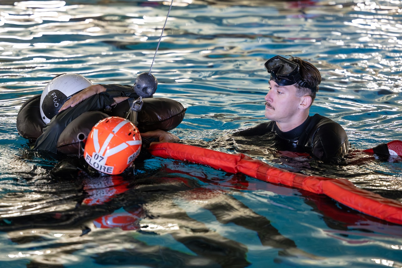 A sailor in a wetsuit instructs another sailor in a wetsuit in a pool.