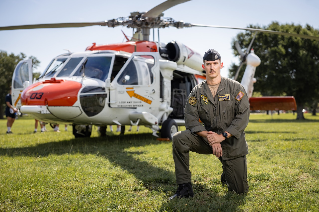 A sailor kneels in grass next to an orange and white helicopter on a sunny day.