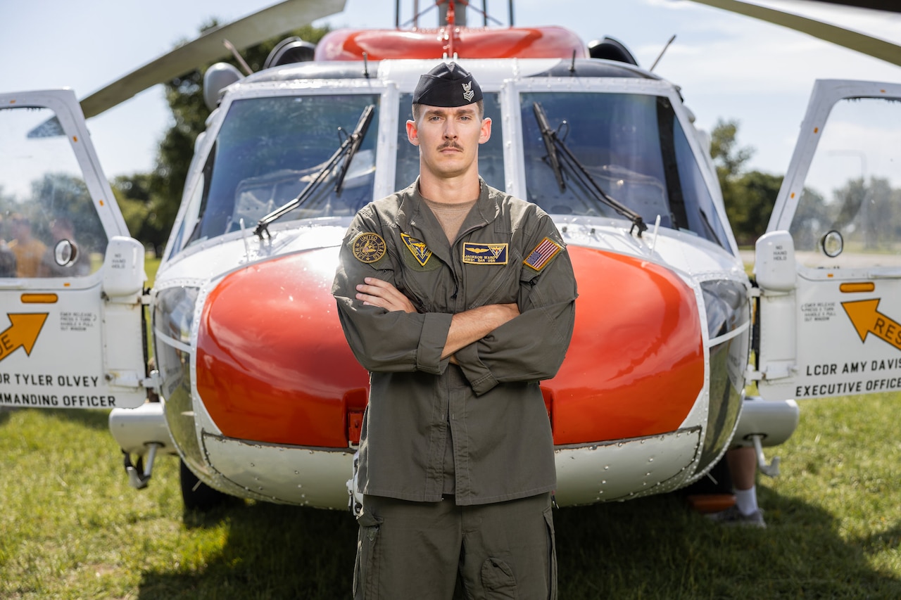 A sailor stands in front of a red and white helicopter parked on a field on a sunny day