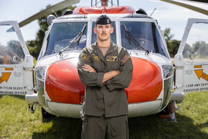 A sailor stands in front of a red and white helicopter parked on a field on a sunny day
