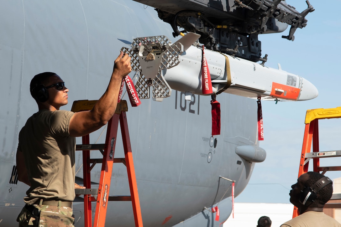 An airman stands on a ladder and adjusts the end of a missile attached to an aircraft as another airman looks on.