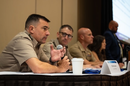 A soldier seated at a table speaks into a microphone. There are two soldiers sitting next to him and two civilians in the background.