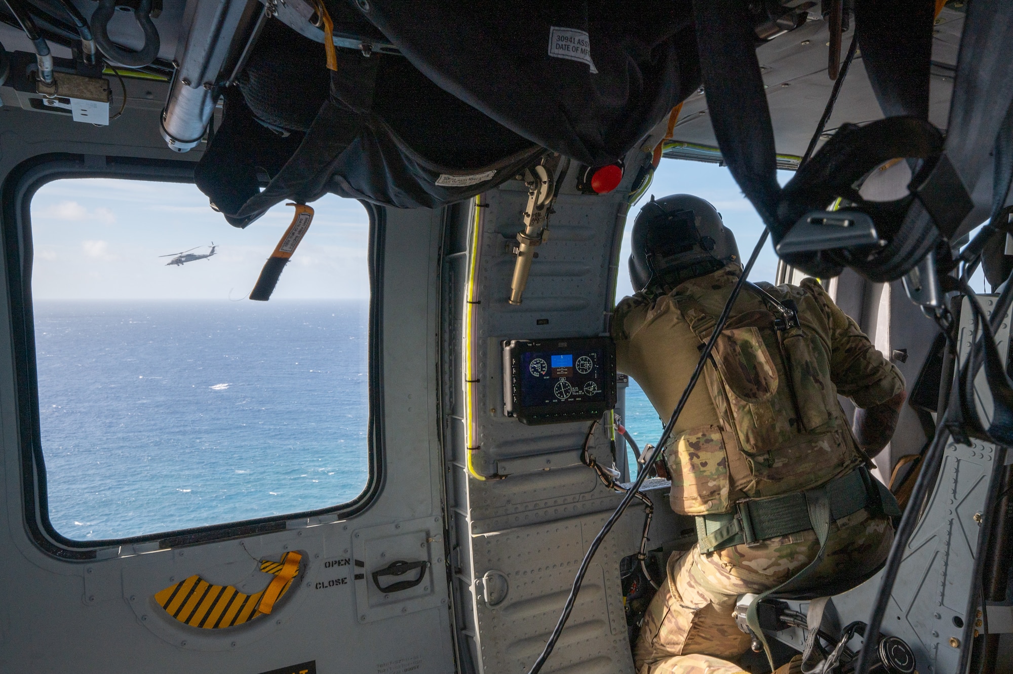 military members looks out of a window inside of a helicopter while another helicopter flies nearby and is seen through the window