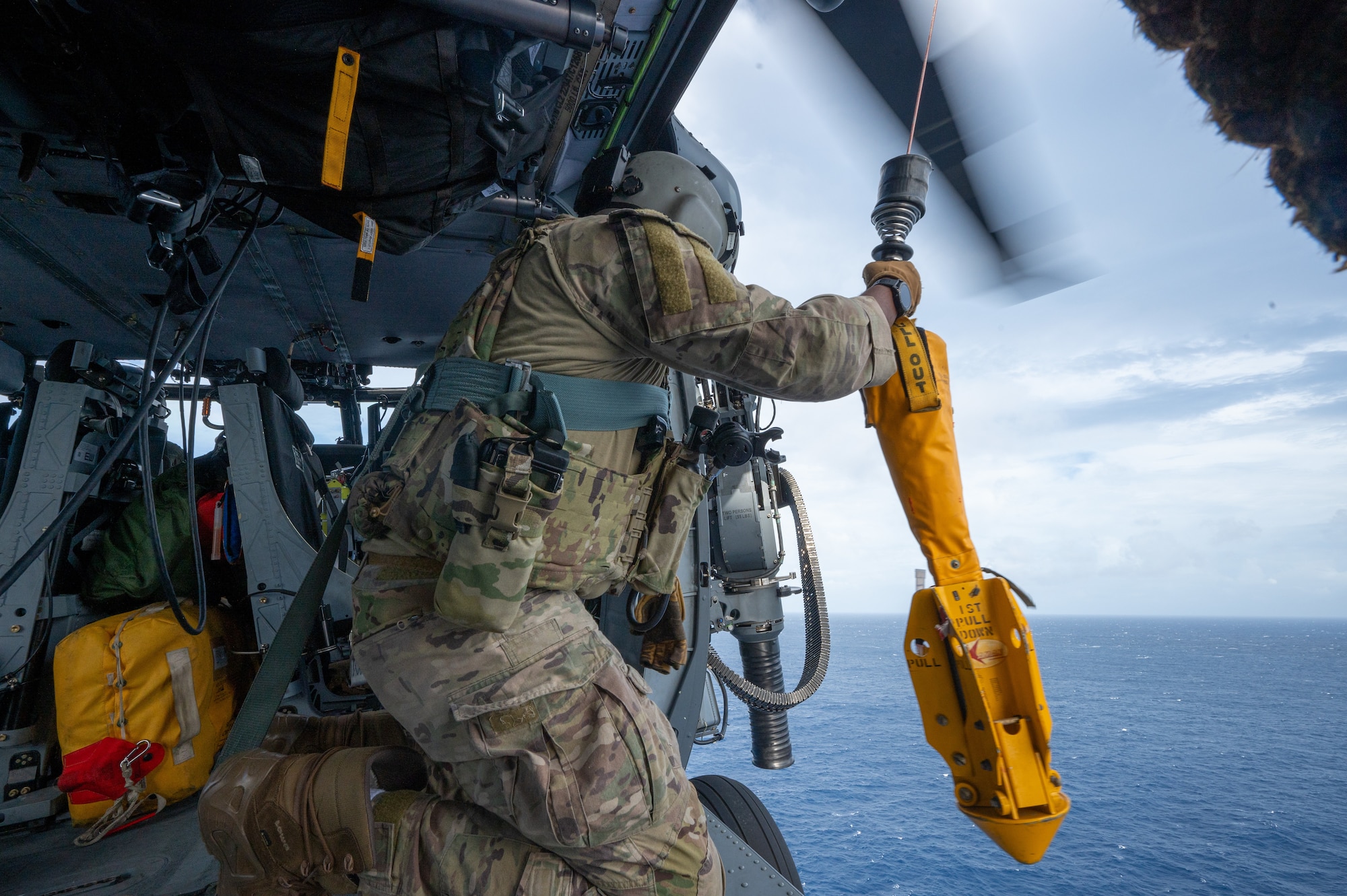 a military member kneels inside of a helicopter while holding a yellow retractible seat attached to a rope hoist