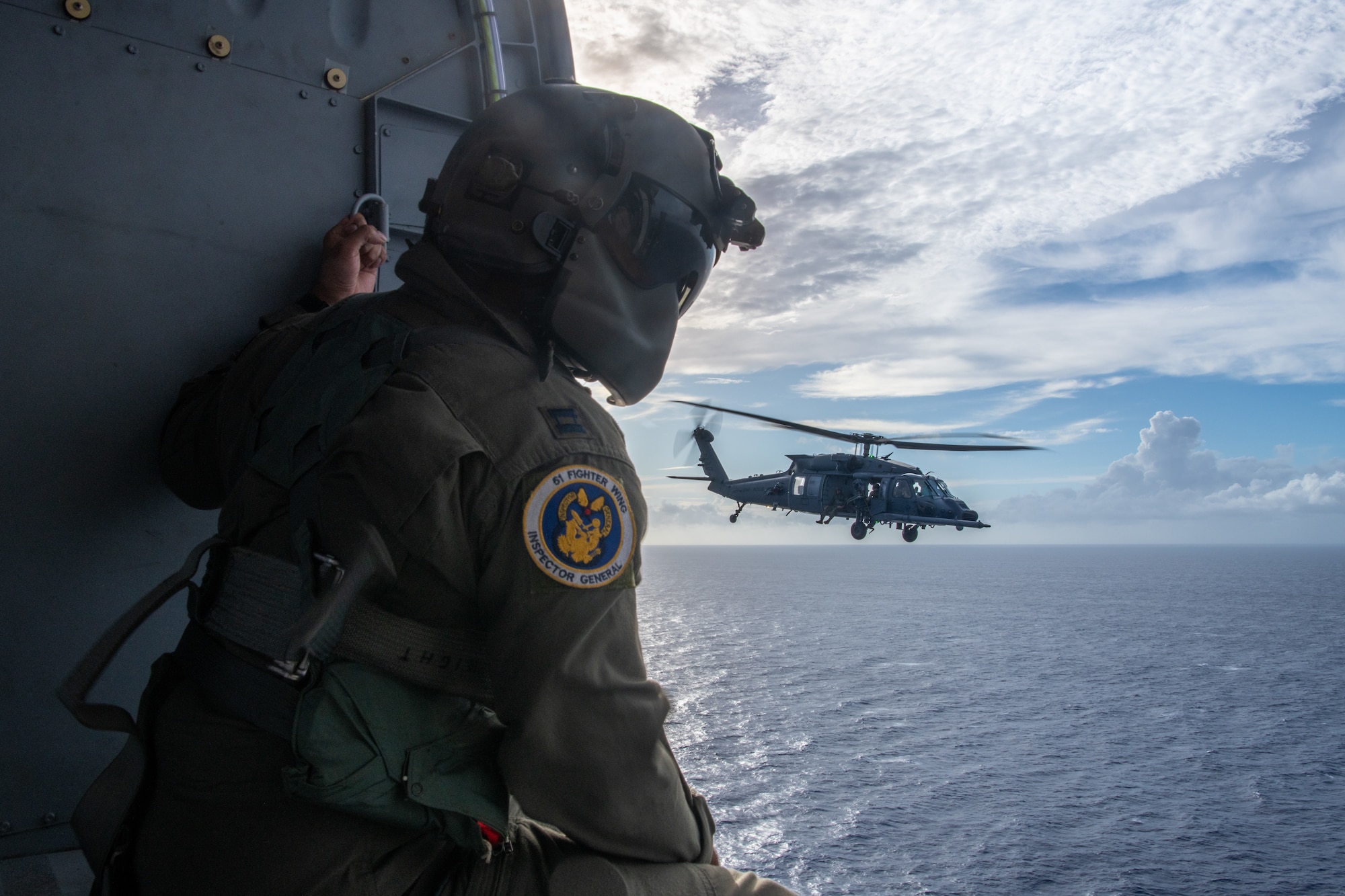 military member sits in a helicopter and looks out to the water with another helicopter flying close by