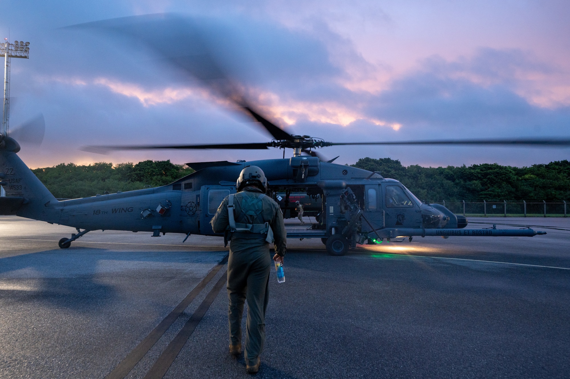 a military member with a pilot helmet walks toward a helicopter as the rotors spin