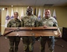 Three Army leaders pose with a large award plaque in front of the United States flag and flags representing the Army Recruiting Battalions of the 3rd Recruiting Brigade