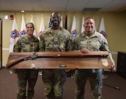 Three Army leaders pose with a large award plaque in front of the United States flag and flags representing the Army Recruiting Battalions of the 3rd Recruiting Brigade