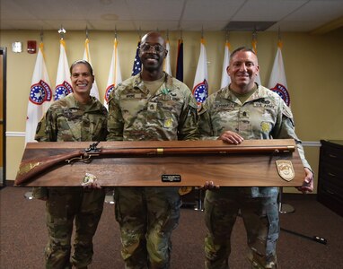 Three Army leaders pose with a large award plaque in front of the United States flag and flags representing the Army Recruiting Battalions of the 3rd Recruiting Brigade