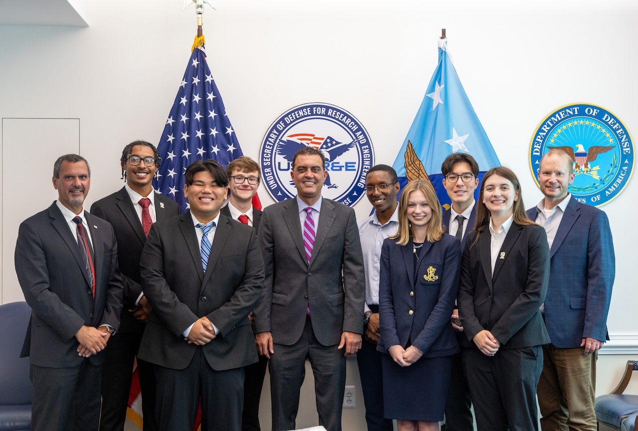 Several men and women in business attire pose for a photo with the American and Defense Department flags in the background.