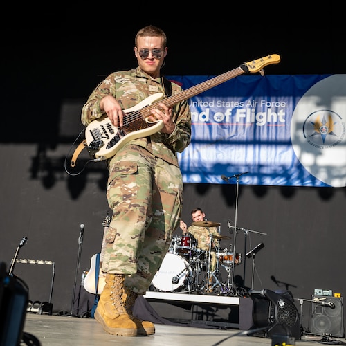 A man, wearing military camouflage uniform and mirrored sunglasses, plays a off-white bass guitar. Another military member can be seen in the background playing a drum set under a blue and white banner reading Band of Flight in white text