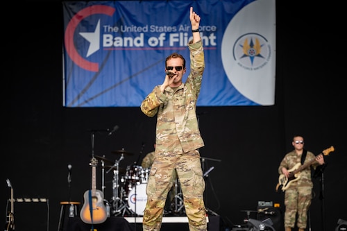 A man, wearing a military camouflage uniform, points a finger to the sky as he holds a microphone to his mouth. Another military member can be seen in the background with a bass guitar, and a blue and white banner that reads Band of Flight in white text