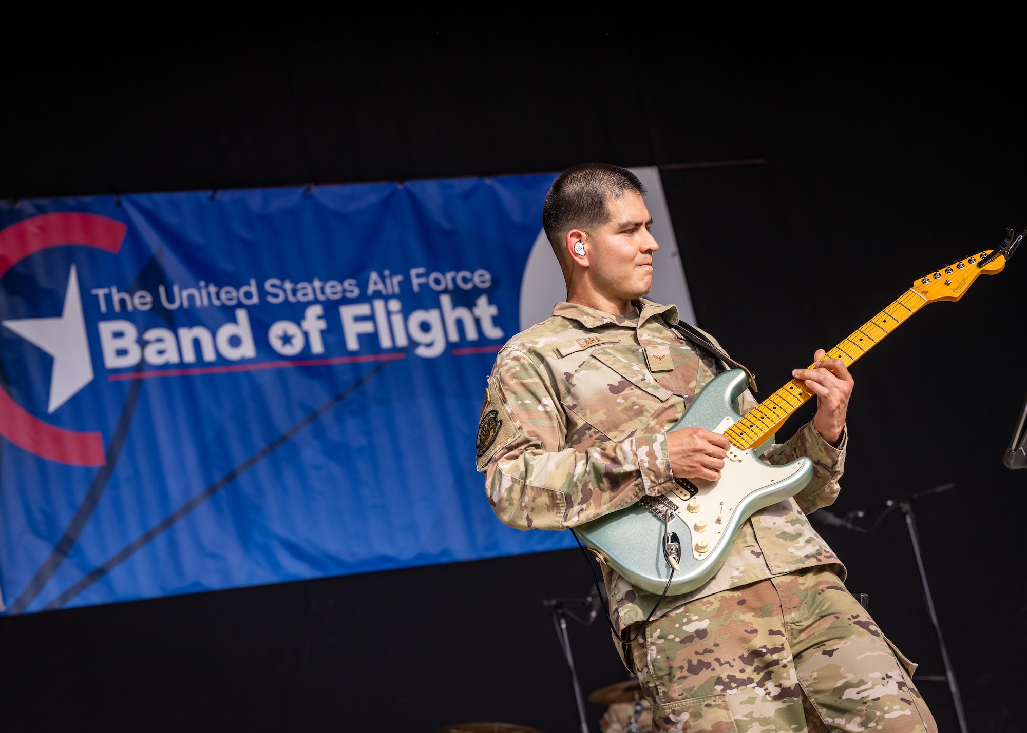 A man, wearing military camouflage uniform, plays a light blue and white guitar in front of a blue banner with white letters reading Band of Flight