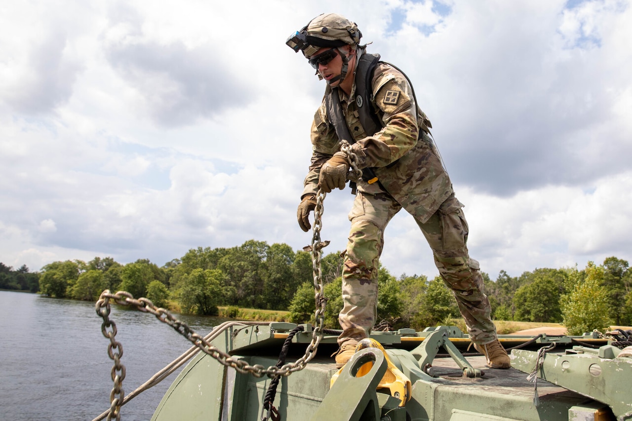 A solider in a camouflage uniform and a helmet removes a chain from a large floating bridge. There is a river and trees in the background.