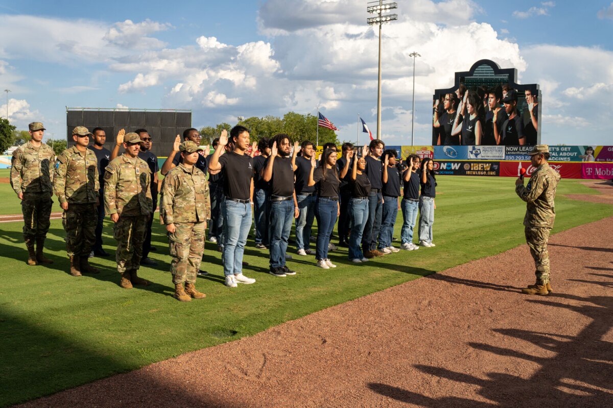 Civilians in black shirts and blue jeans raise their right hand as troops in formation stand at attention while a soldier raising his right hand stands in front of the formation.