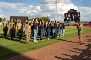 Civilians in black shirts and blue jeans raise their right hand as troops in formation stand at attention while a soldier raising his right hand stands in front of the formation.