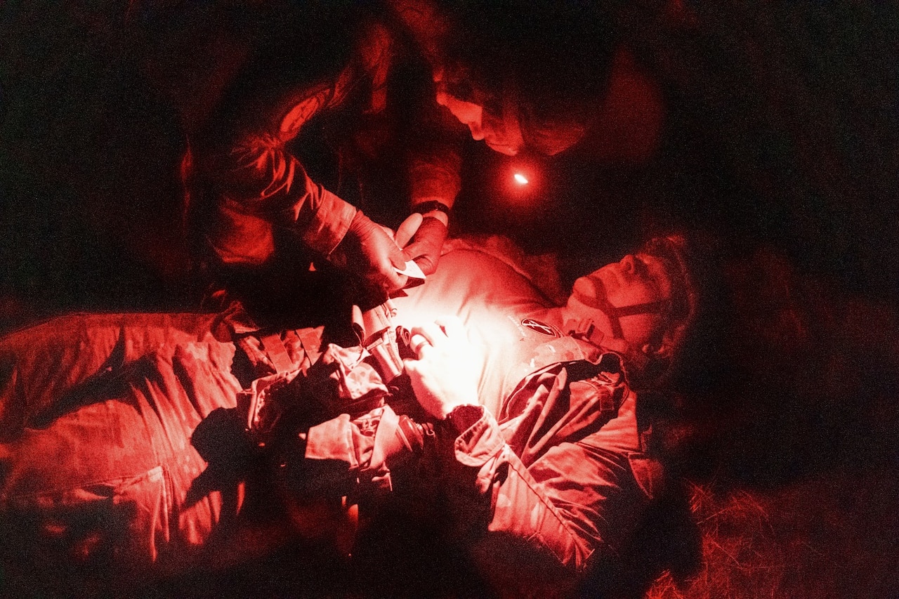 A soldier in a camouflage uniform and a helmet lies on the ground as another soldier in similar attire supplies medical care.
