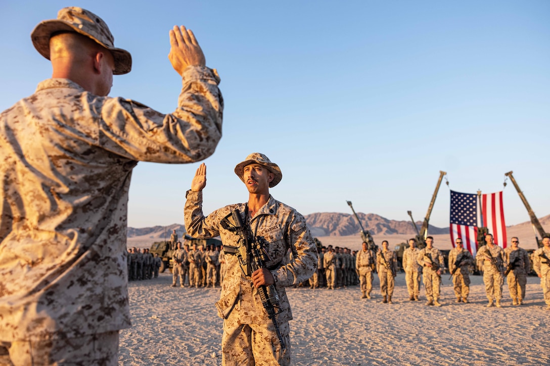 Two Marines face each other while raising their right hands in a desert-like area as dozens service members in formation watch in front of an American flag.