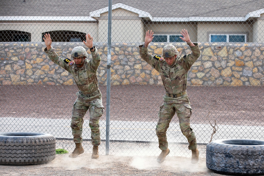 Two soldiers in tactical gear jump in between tires in front of a fence.