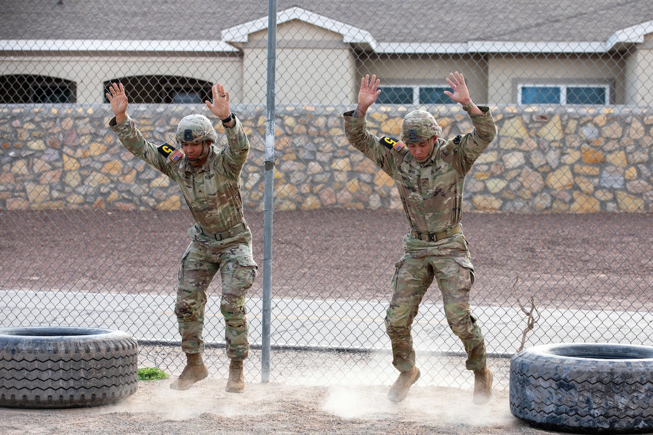 Two soldiers in tactical gear jump in between tires in front of a fence.