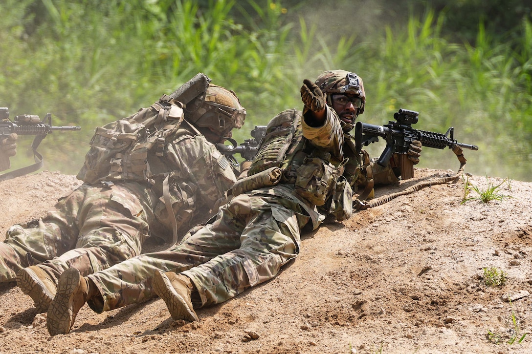 A soldier laying in the dirt next to a fellow soldier holding a weapon looks back raising their hand.