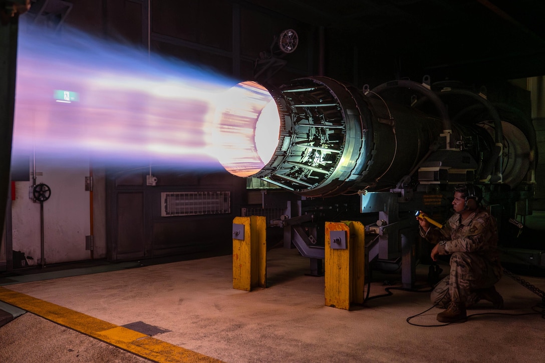 An airman using a flashlight kneels to inspect an aircraft’s engine as it creates a blue, white and orangish glow.