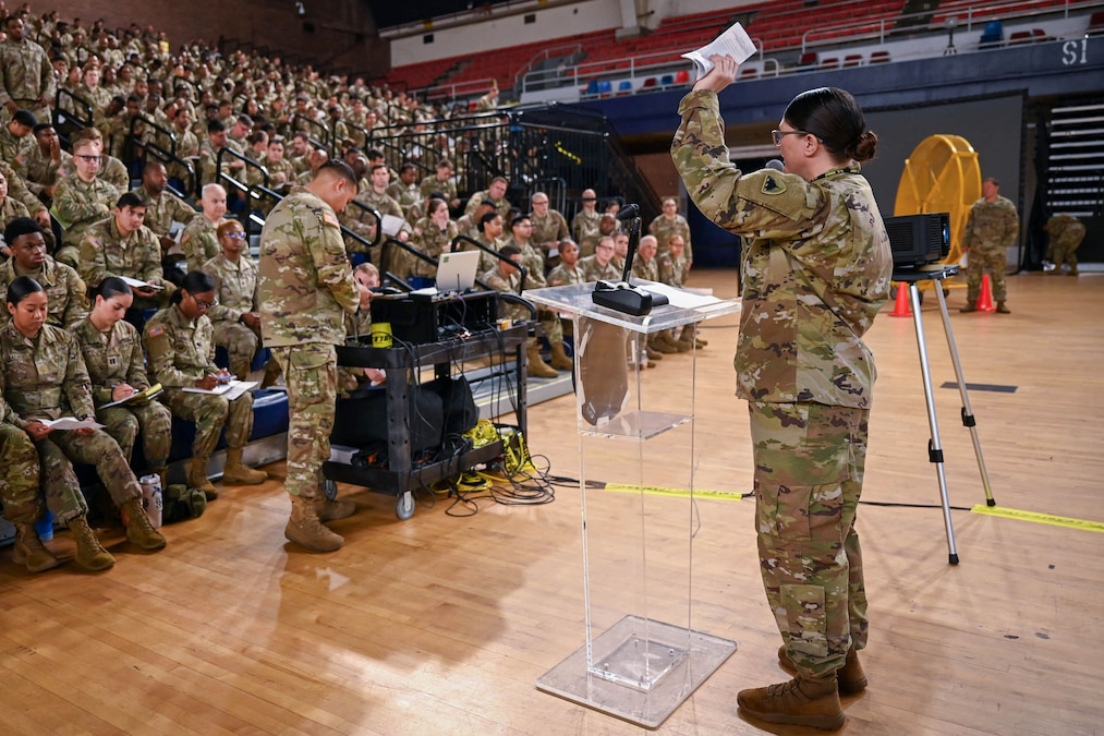 A soldier speaks to dozens of guardsmen sitting in bleachers in a gymnasium.