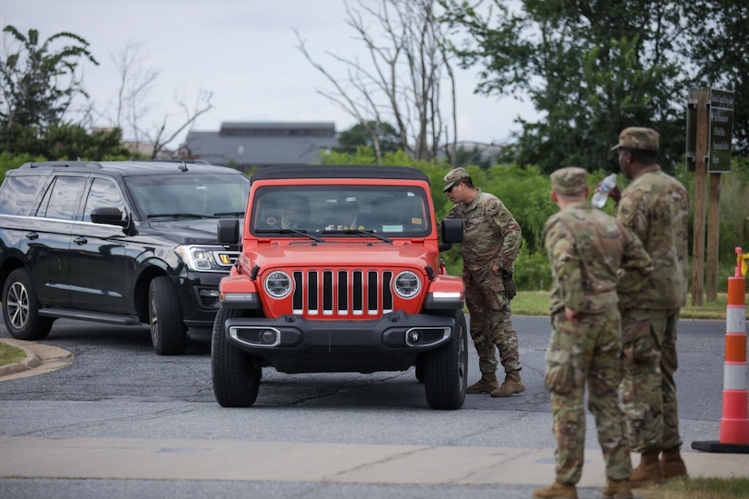 A military member stands at the driver's side of a red jeep and talks with the driver. Other servicemembers watch.