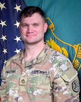Man in U.S. Army uniform standing in front of two flags.