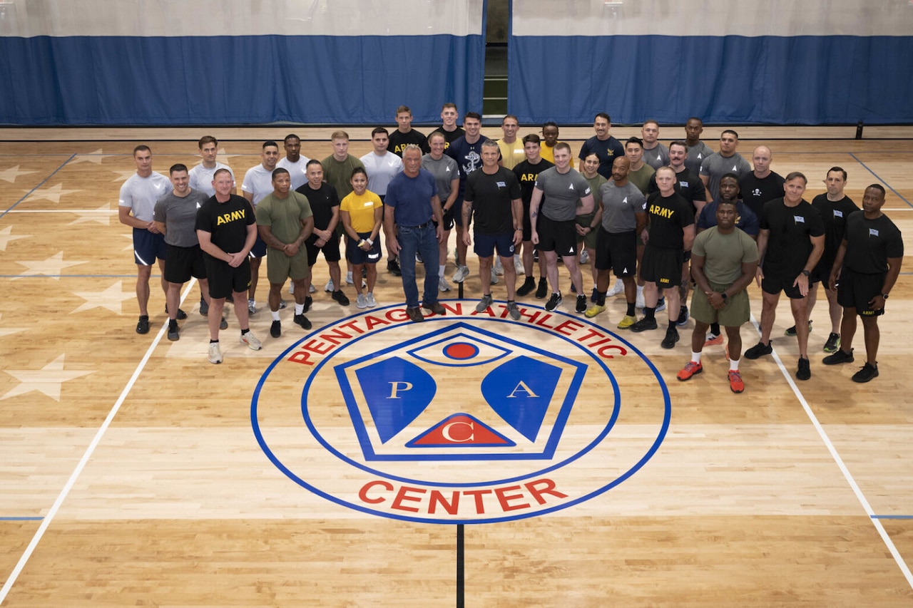 Roughly three dozen men and women wearing athletic attire – and one man in a tee shirt in jeans – are in formation posing for a group photo on a wooden basketball court with a large, circular logo on the floor that reads “Pentagon Athletic Center.”