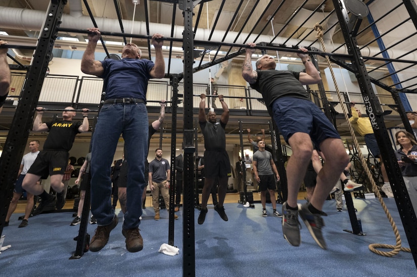 Five men in athletic gear are doing pull-ups in an indoor gymnasium as other people in the background observe them.
