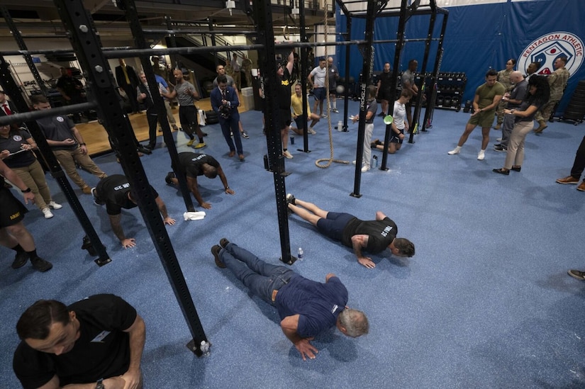 A group of men in athletic gear and one additional man in a tee shirt and jeans are performing push-ups on the floor of an indoor gymnasium while others observe them.