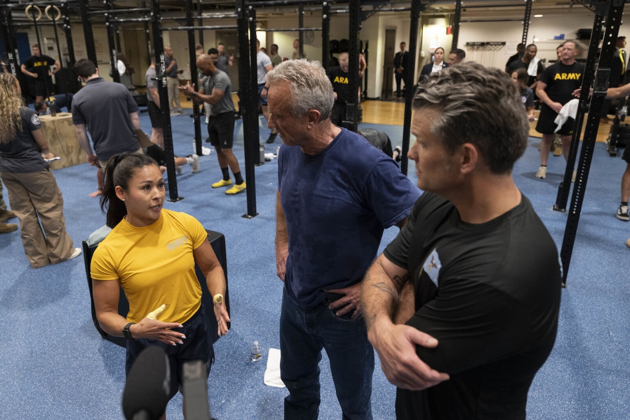 Three people, a man and a woman in athletic attire and a second man in a tee shirt and jeans, are having a conversation in an indoor gymnasium.