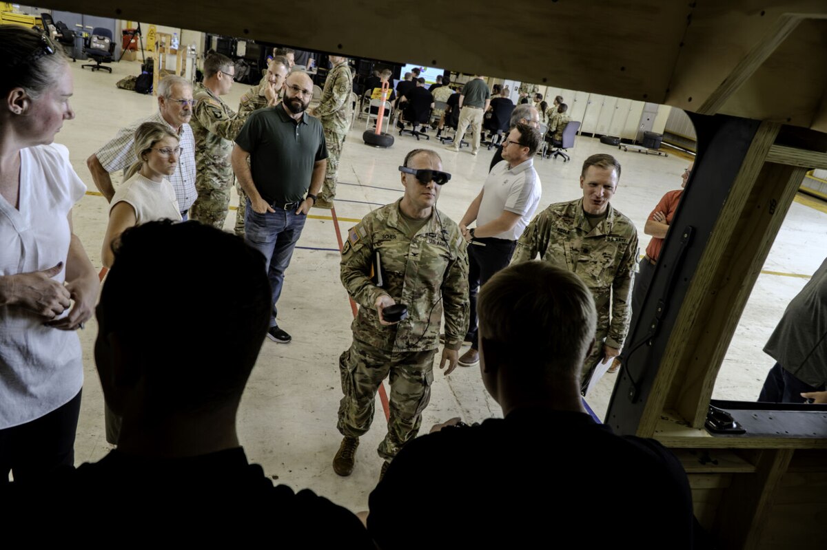 A soldier in a camouflage uniform wearing virtual reality goggles is surrounded by several people in casual attire and another soldier in a camouflage uniform.