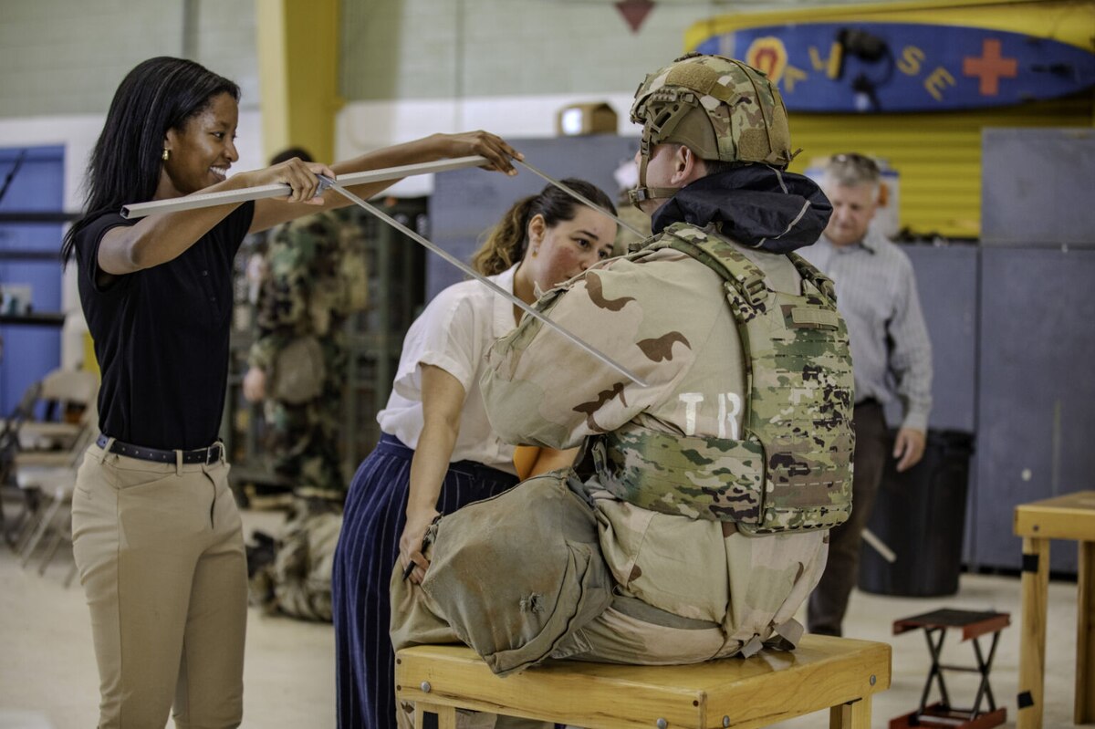 A woman in business casual attire holds a measuring device up to a soldier wearing a camouflage uniform who is sitting in front of her.
