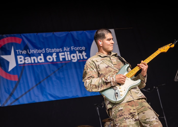 A man, wearing military camouflage uniform, plays a light blue and white guitar in front of a blue banner with white letters reading Band of Flight