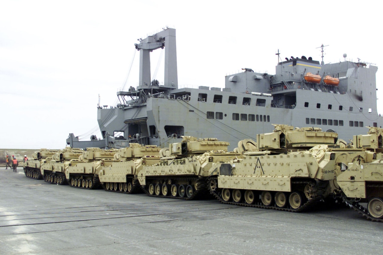 Military vehicles line up in a row in front of a cargo ship.
