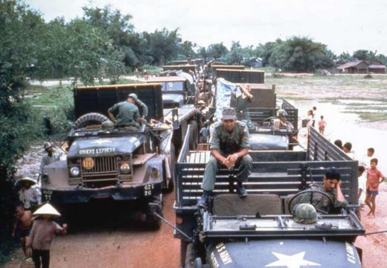 Several flatbed trucks are lined up along a dirt road. People in military uniforms are seated in and atop the trucks, while others stand alongside them.