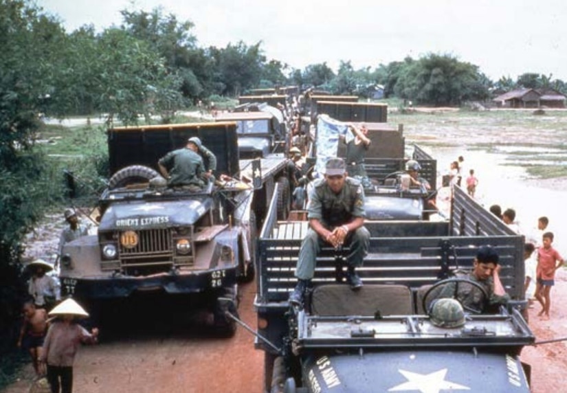 Several flatbed trucks are lined up along a dirt road. People in military uniforms are seated in and atop the trucks, while others stand alongside them.
