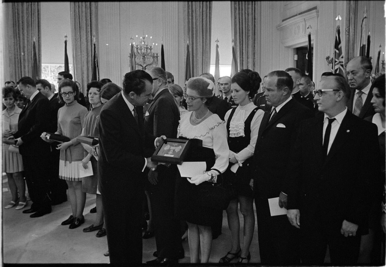 A man wearing business attire hands a woman a small plaque. Several people stand behind the woman, watching.