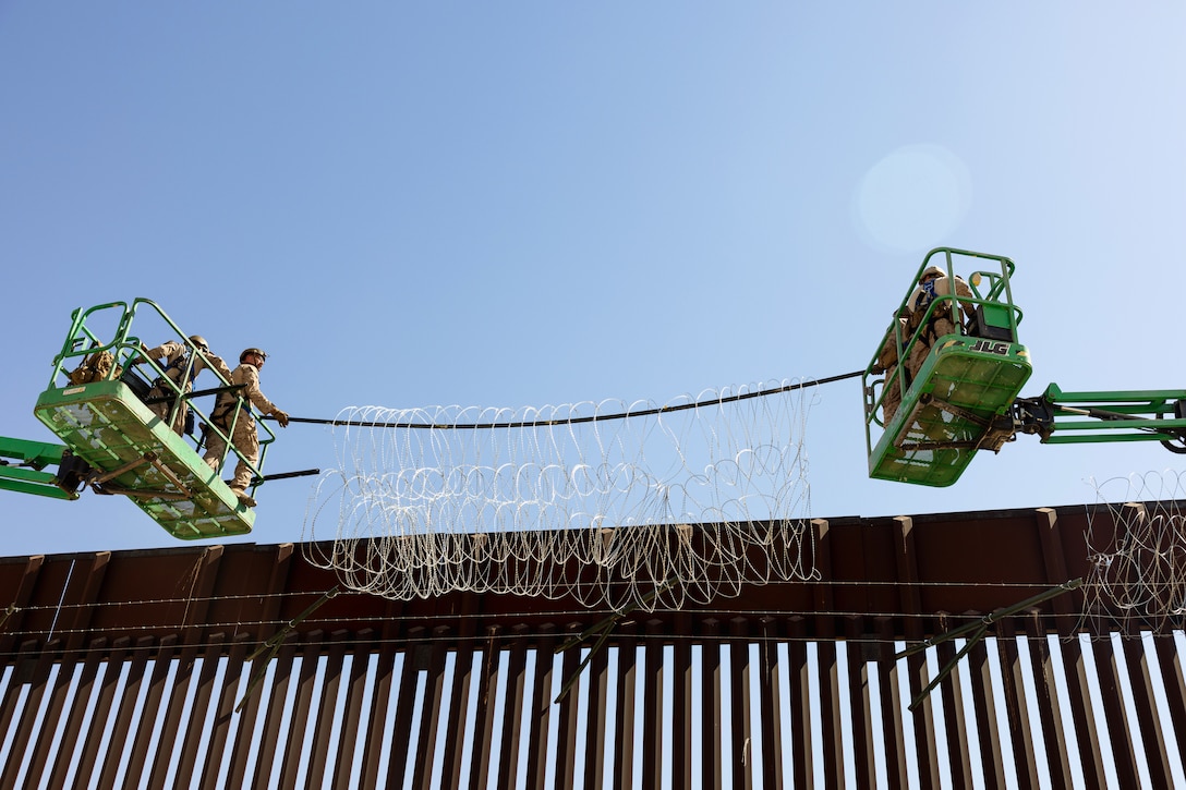 Marines hold out concertina wire from raised platforms at the top of a metal wall under a blue sky.