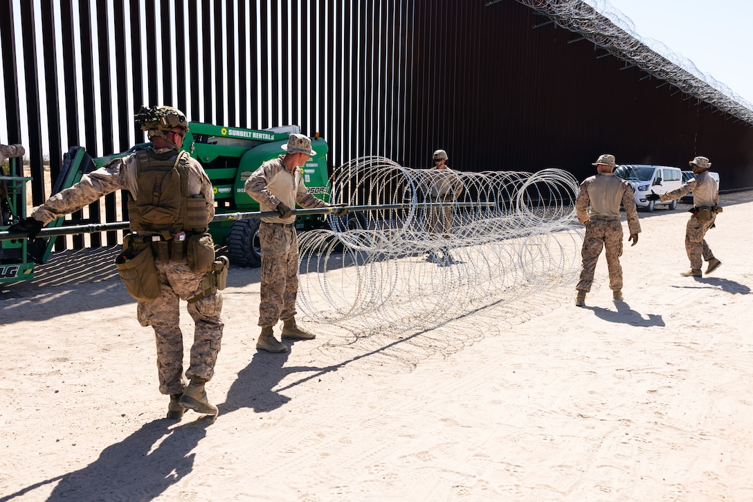 Marines hold concertina wire next to a tall wall on a dirt road on a sunny day.