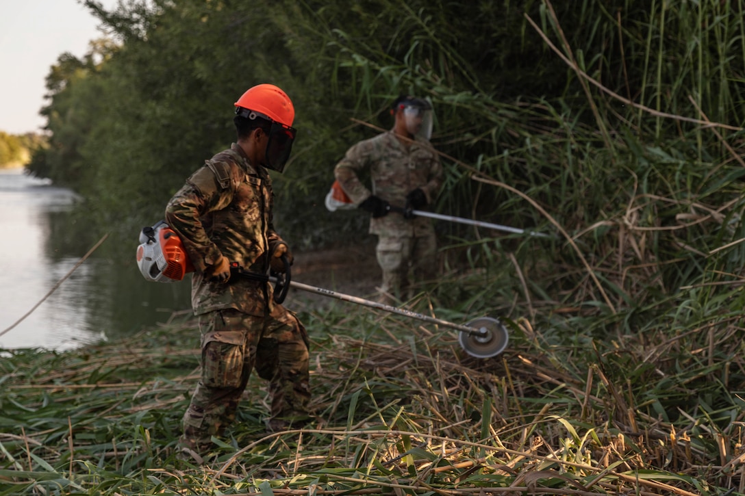 Two soldiers wearing helmets hold string trimmers and clear vegetation next to a river.