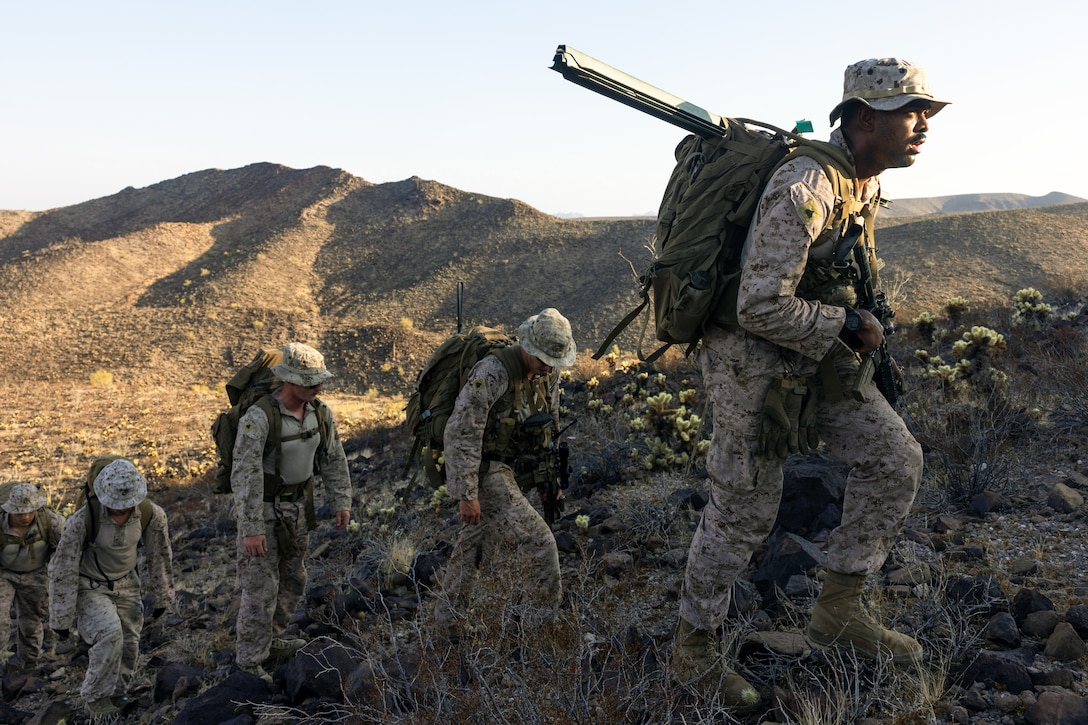 Five Marines walk in a line up a grassy hill with a shadowed ridge in the background on a sunny day.