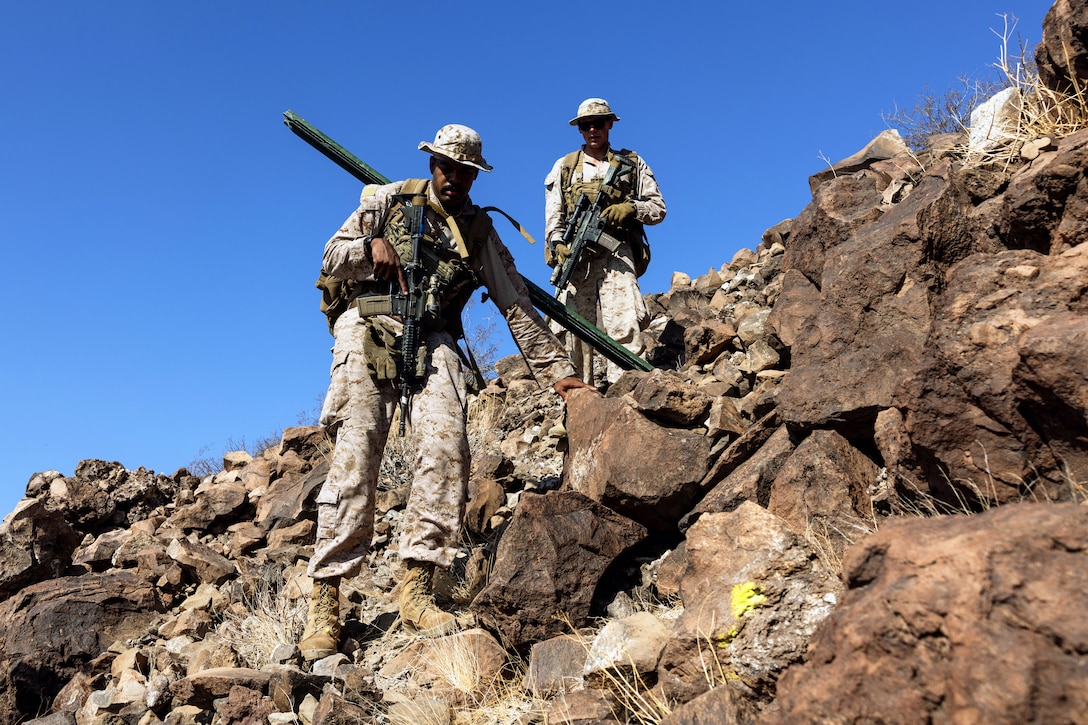Two Marines stand on a rocky outcrop with gear and a sign under a blue sky.