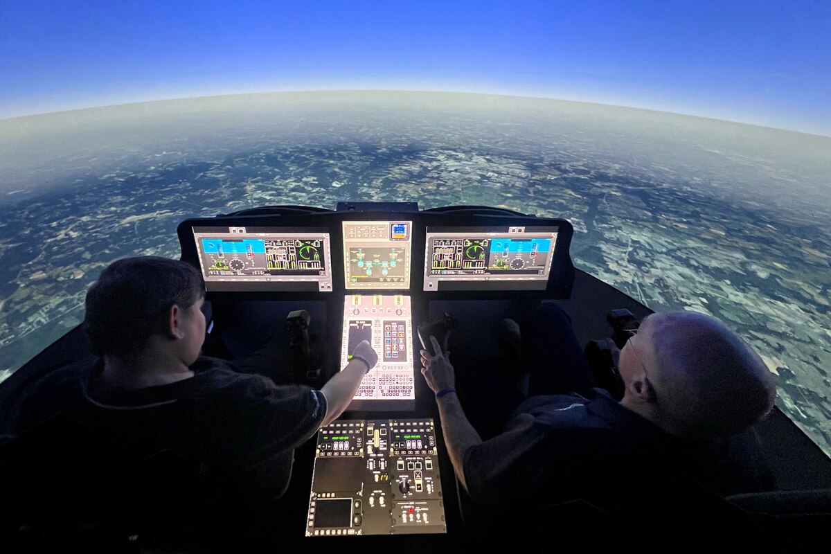 Two people sit in simulated cockpits with two screens and a console in the middle with a projection of the sky in front.